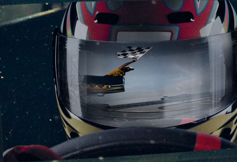 A close-up of a racecar driver with two hands on the wheel and a reflection of a grand marshall waving the checkered winner's flag. 